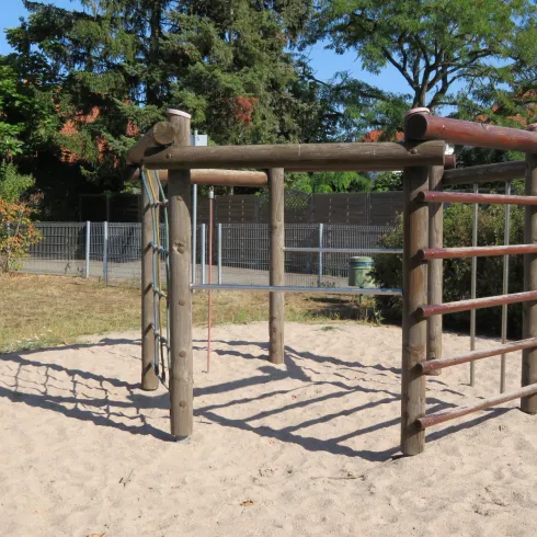 Ein Spielplatz mit einer Holzklettergerüst steht auf einem Sandboden. Im Hintergrund sind Bäume und ein Zaun zu sehen.