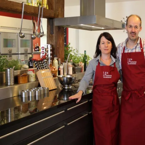 Ein modernes Küchenatelier mit zwei Personen in roten Schürzen. Die Küche ist gut ausgestattet, mit Pflanzen und Kochutensilien im Hintergrund.
