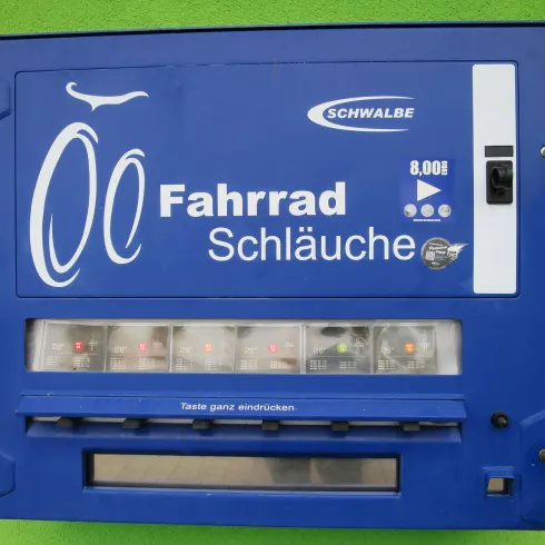 Ein blauer Automaten für Fahrradschläuche an einer grünen Wand. Der Automat bietet verschiedene Schläuche zum Verkauf an.