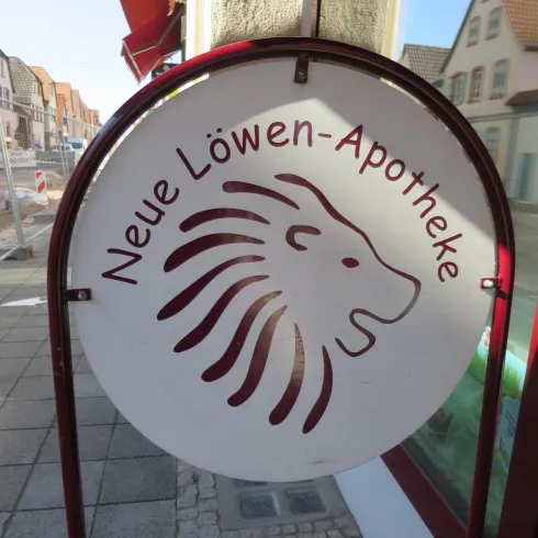 Ein Schild der "Neuen Löwen-Apotheke" mit einem stilisierten Löwenkopf. Die Apotheke ist an einer Straße sichtbar.