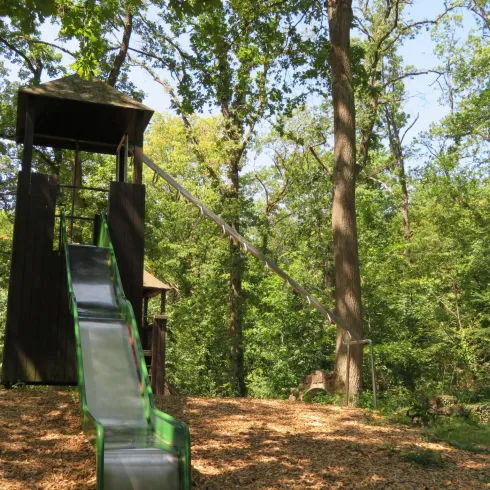 Ein Spielplatz im Wald mit einer Rutsche und einem Spielturm. Umgeben von Bäumen und Natur.
