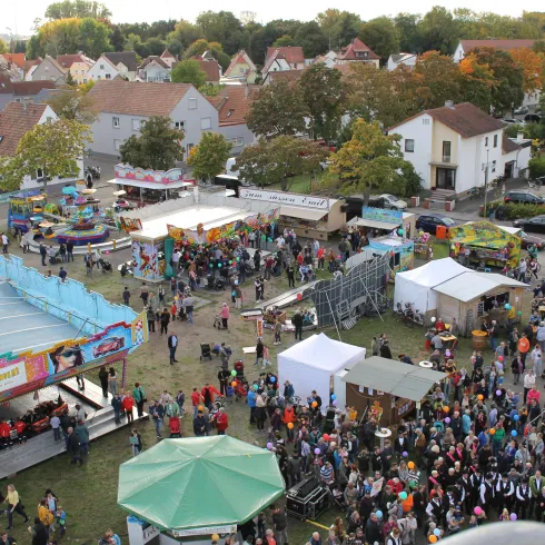 Eine lebendige Veranstaltung mit vielen Menschen und verschiedenen Ständen. Im Vordergrund sind Fahrgeschäfte und Verkaufsstände zu sehen, umgeben von bunten Bäumen im Herbst.