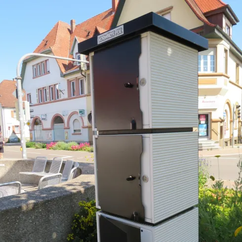Ein moderner Briefkasten mit mehreren Fächern steht an einer Straßenecke. Im Hintergrund sind Gebäude und eine kleine Grünfläche zu sehen.