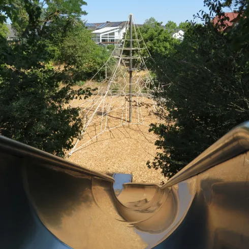 Eine Rutsche mit Blick auf einen Kletterturm aus Seilen auf einem Spielplatz. Der Bereich ist von Bäumen umgeben und hat einen Kiesboden.