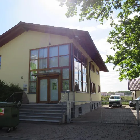 Ein modernes Gebäude mit großen Fenstern und einem einladenden Eingangsbereich. Im Hintergrund sind Bäume und ein parkendes Auto zu sehen.