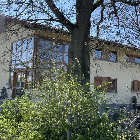 Ein modernes Haus - Forstamt - mit großen Fenstern und einem begrünten Außenbereich. Bäume und Pflanzen umgeben die Gebäude.