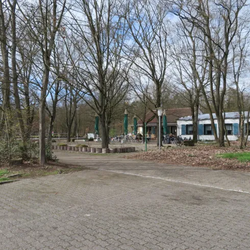 Ein Parkplatz mit kahlen Bäumen und einem gepflasterten Weg. Im Hintergrund sieht man ein Gebäude mit einer Terrasse und Fahrrädern.