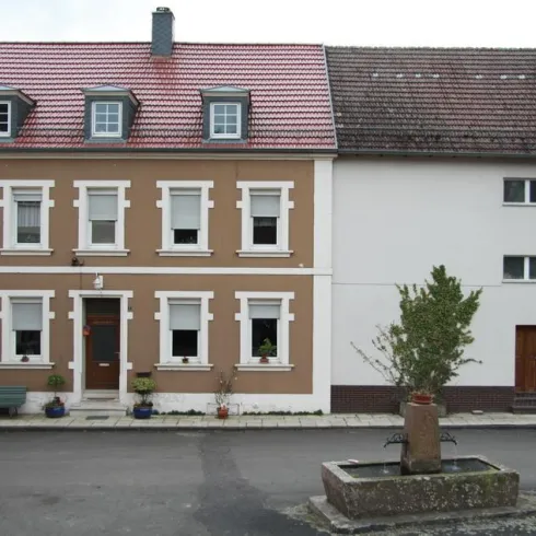 Ein zweigeteiltes Gebäude mit einem braunen und einem weißen Teil. Im Vordergrund befindet sich ein Brunnen und einige Pflanzen.