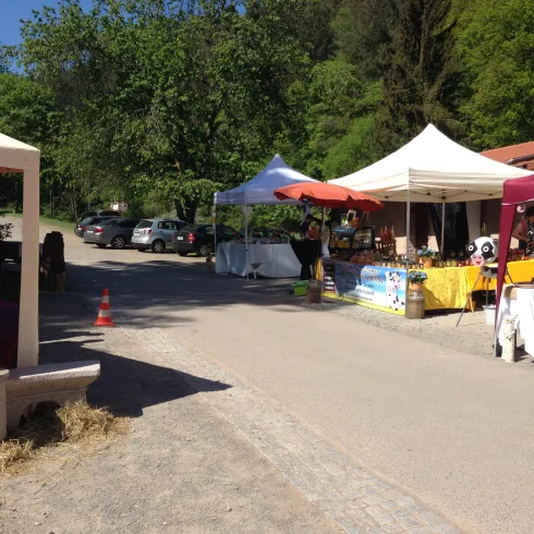 Ein kleiner Markt mit unterschiedlichen Ständen und Zelten an einer Straße. Im Hintergrund sind Bäume und eine ländliche Umgebung zu sehen.