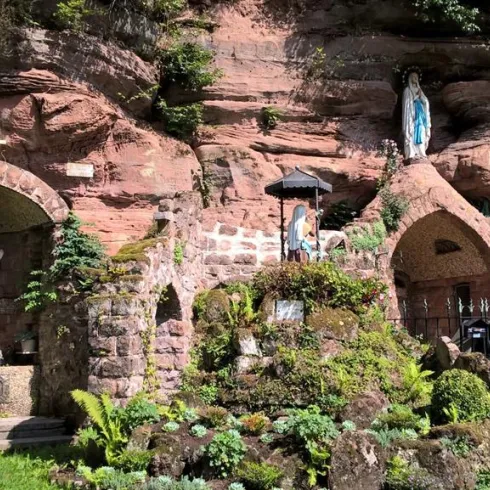 Eine kleine Kapelle in einer Felswand, umgeben von üppigem Grün. Im Vordergrund sind Statuen und eine dekorative Gestaltung sichtbar.