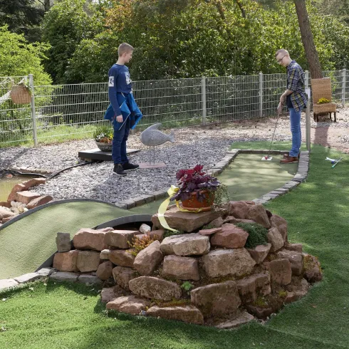 Zwei Männer spielen Minigolf in einem Garten. Der Platz ist mit Gras, Steinen und einem kleinen Teich gestaltet.