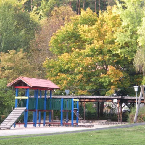 Ein Spielplatz mit bunten Geräten steht in einem Park. Im Hintergrund sind große, grüne Bäume zu sehen.