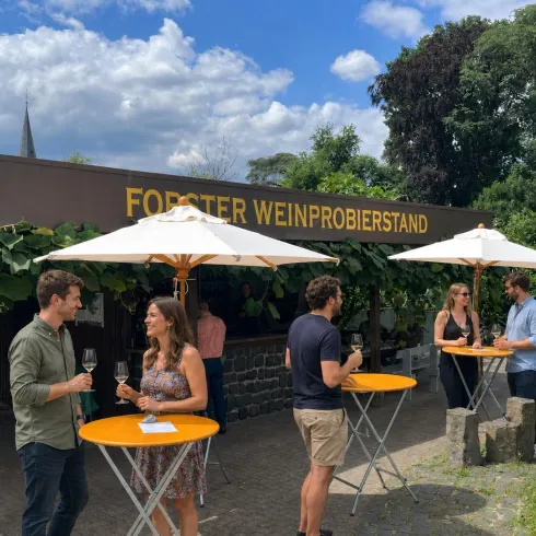 Eine Weinprobe im Freien mit mehreren Tischen und Sonnenschirmen. Menschen genießen den Wein und die entspannte Atmosphäre in einem schönen Garten.