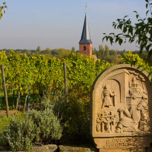 Eine steinerne Reliefskulptur steht inmitten von Weinreben. Im Hintergrund sieht man einen Kirchturm und eine ländliche Landschaft.