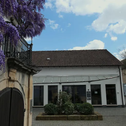 Ein historisches Gebäude mit modernen Fenstern und einem schönen Garten. Im Vordergrund blühen lila Pflanzen und der Himmel ist teilweise bewölkt.