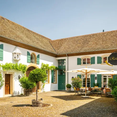 Ein einladender Innenhof mit grünen Fensterläden und Pflanzen. Tische und Stühle laden zum Entspannen unter einem Sonnenschirm ein.