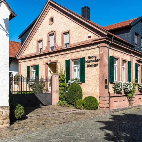 Ein charmantes Weingut mit einer roten Backsteinfassade und grünen Fensterläden. Die Umgebung ist gepflegt mit Büschen und einem ruhigen Straßenverlauf.