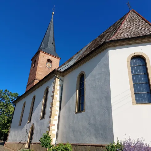 Eine Kirche mit einem hohen, spitzen Turm und einem schlichten weißen Äußeren. Der Himmel ist klar und blau, umgeben von Bäumen und Grünflächen.