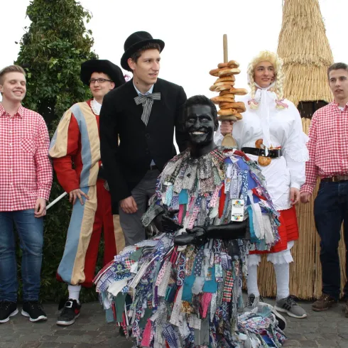 Eine Gruppe von Menschen in traditionellen Kostümen steht auf einer Feier. Im Vordergrund sitzt eine Person in einem bunten kostümierten Outfit.