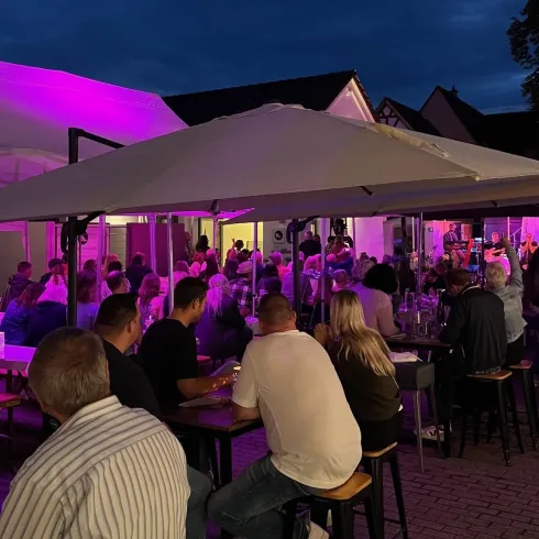 Ein belebtes Restaurant mit vielen Gästen unter Sonnenschirmen. Die Umgebung ist Abenddämmerung mit bunten Lichtern.