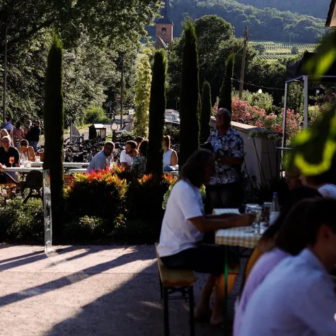 Eine lebhafte Außenrestaurant-Szenerie mit Gästen, die an Tischen sitzen und essen. Im Hintergrund sind Weinberge und Bäume zu sehen.