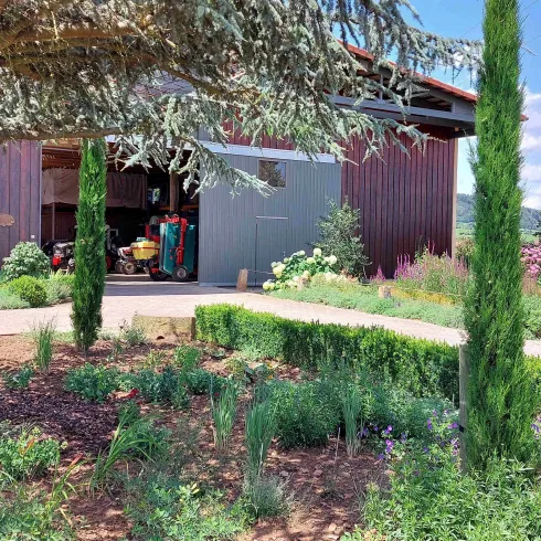 Ein schöner Garten mit bunten Pflanzen und hohen Zypressen. Im Hintergrund steht eine Holzscheune unter einem blauen Himmel.