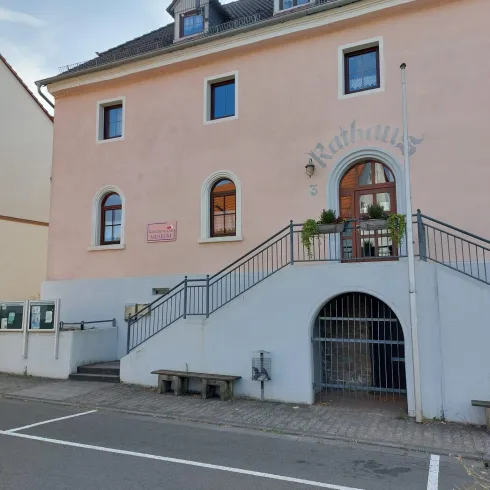 Ein modernes Rathaus mit einer hellrosa Fassade und mehreren Fenstern. Die Eingangstreppe ist von Pflanzen umgeben und es gibt eine Straßensignalisierung vor dem Gebäude.