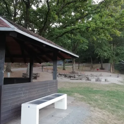 Ein überdachter Platz mit einer Bank steht in einem ruhigen Bereich. Im Hintergrund sind mehrere Picknicktische und Bäume zu sehen.