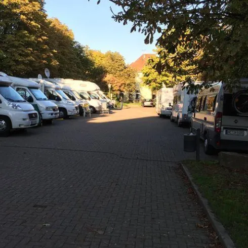 Ein Parkplatz mit mehreren Wohnmobilen, die in einer Reihe geparkt sind. Die Umgebung ist von Bäumen umgeben und es ist ein klarer Himmel sichtbar.