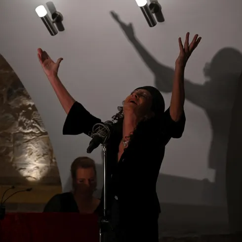 Eine Sängerin steht auf der Bühne und hebt ihre Arme ausdrucksvoll. Im Hintergrund ist ein Musiker am Klavier sichtbar.