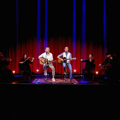 Zwei Musiker spielen live auf der Bühne Gitarre und singen. Im Hintergrund stehen weitere Musiker mit Streichinstrumenten.