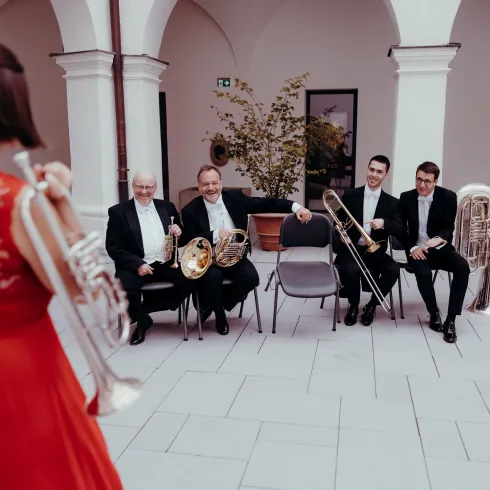 Eine Musikerin in einem roten Kleid spielt vor einer Band. Im Hintergrund sitzen vier Musiker mit verschiedenen Blasinstrumenten.