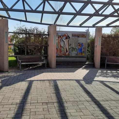 Ein ruhiger Platz mit Sitzbänken unter einem Gestell aus Glas und Metall. Im Hintergrund sind Bäume und eine Wand mit Kunstwerken zu sehen.