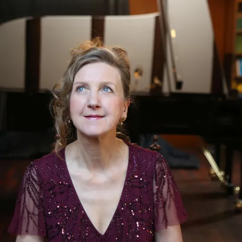 Eine Frau mit einem freundlichen Ausdruck sitzt vor einem Klavier. Der Raum hat eine elegante Atmosphäre mit Bücherregalen im Hintergrund.
