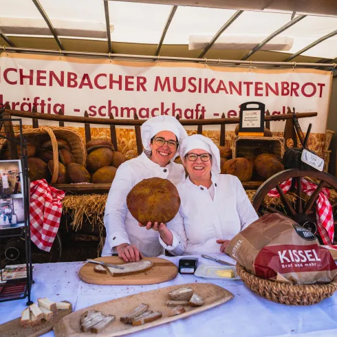 Zwei Bäckerinnen in weißen Schürzen präsentieren frisch gebackenes Brot an einem Marktstand. Im Hintergrund sind weitere Brote und eine bunte Verkaufsdekoration zu sehen.