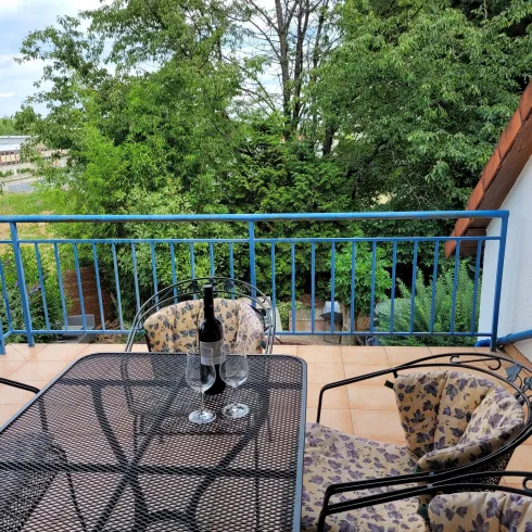 Ein schöner Balkon mit einem Tisch und Stühlen aus schwarzem Metall. Auf dem Tisch stehen zwei Gläser und eine Flasche, umgeben von grünen Bäumen.