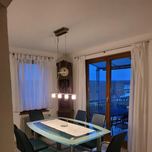 Ein gemütlicher Essbereich mit einem Glas-Tisch und sechs Stühlen. Große Fenster lassen Licht herein und bieten einen Blick nach draußen. Über dem Tisch hängt eine Pendelleuchte. An den Fenster hängen weiße Vorhänge.