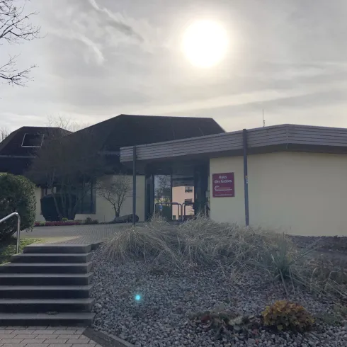 Ein modernes Gebäude mit einem glatten Eingang und einer Treppe, umgeben von Pflanzen. Die Sonne scheint leicht über dem Dach.