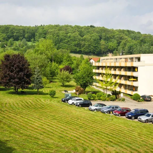 Eine ruhige Landschaft mit einem Hotel und vielen Bäumen. Auf dem Parkplatz sind mehrere Autos geparkt.
