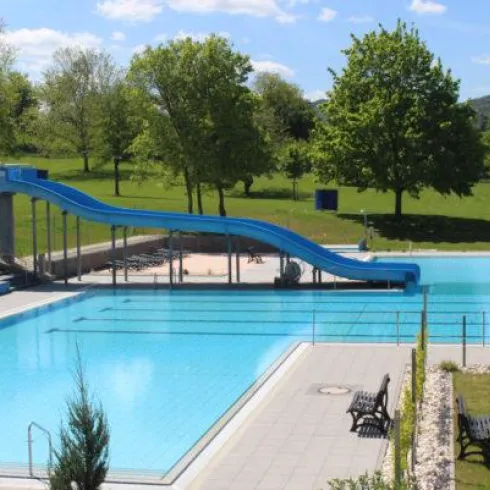 Ein Schwimmbad mit einer blauen Rutsche und klarem Wasser. Umgeben von grünen Bäumen und einer sonnigen Landschaft.