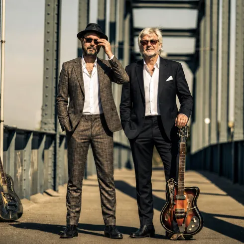 Zwei Männer stehen auf einer Brücke, jeweils elegant gekleidet. Hinter ihnen liegen Gitarren auf dem Boden.