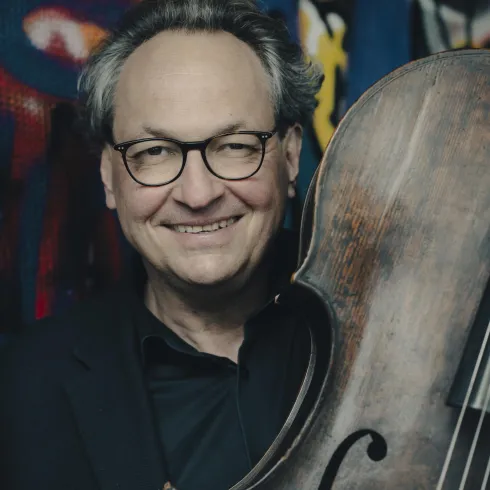 Ein lächelnder Mann hält ein Cello in den Händen. Im Hintergrund sind bunte Graffiti zu sehen.