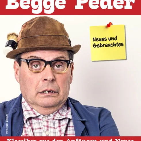 Ein humorvoller Mann mit Brille und Anzug steht vor einem roten Hintergrund. Auf dem Bild ist ein Zettel mit der Aufschrift „Neues und Gebrauchtes“ zu sehen.
