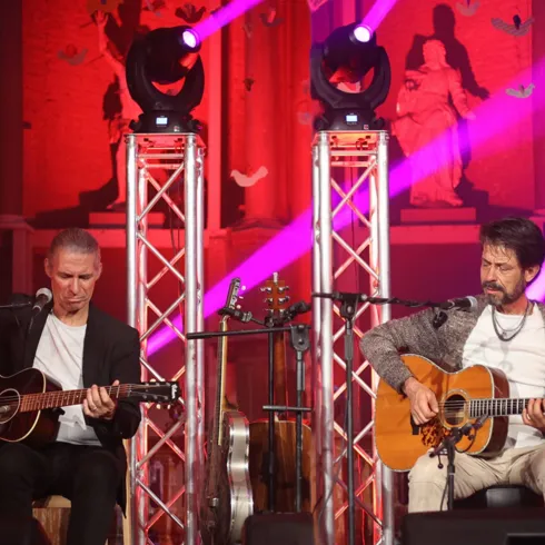 Zwei Musiker spielen akustische Gitarren auf einer Bühne. Bunte Lichter strahlen im Hintergrund und schaffen eine festliche Atmosphäre.