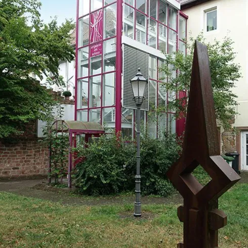 Ein modernes, pinkes Gebäude mit Glasfassade steht in einem Garten. Vor dem Gebäude befindet sich eine auffällige Skulptur und ein Laternenpfahl.