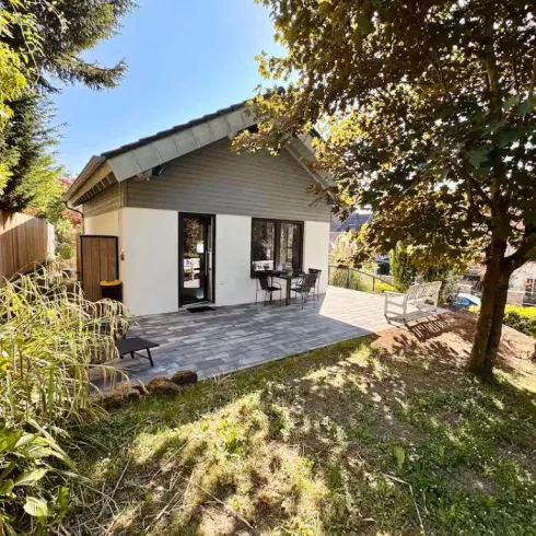 Ein modernes Häuschen umgeben von grünem Gras und Bäumen. Eine Terrasse mit Stühlen lädt zum Entspannen ein.