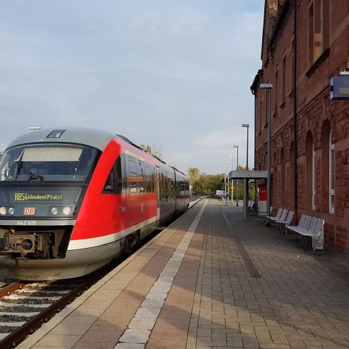 Ein roter Zug steht an einem Bahnhof auf dem Bahnsteig. Die Umgebung ist leicht bewölkt und der Bahnhof ist aus Ziegelstein gebaut.