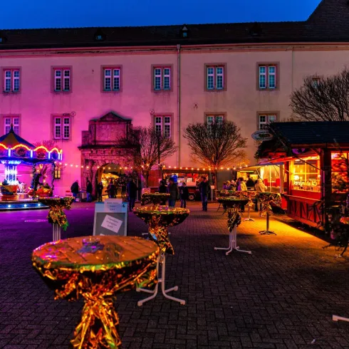 Ein festlich geschmückter Platz mit bunten Lichtern und Ständen. Im Hintergrund sind historische Gebäude zu sehen, während Menschen die Atmosphäre genießen.
