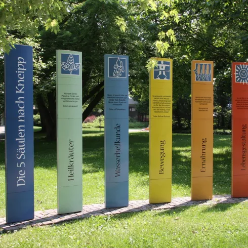 Sechs farbige Säulen stehen in einem Park und repräsentieren die fünf Säulen nach Kneipp. Sie sind umgeben von grünem Grass und Bäumen.