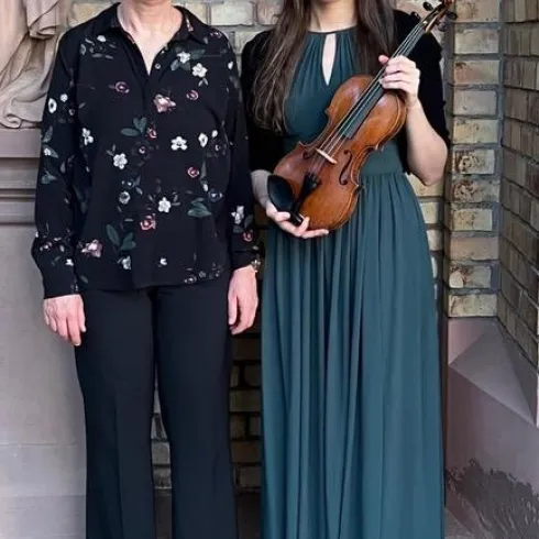 Zwei Frauen stehen vor einer Backsteinmauer. Die jüngere Frau trägt ein elegantes langes Kleid und hält eine Geige in der Hand.